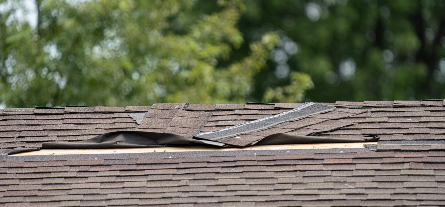 Damaged asphalt shingle roof with lifted and missing shingles exposing the underlayment after storm impact.