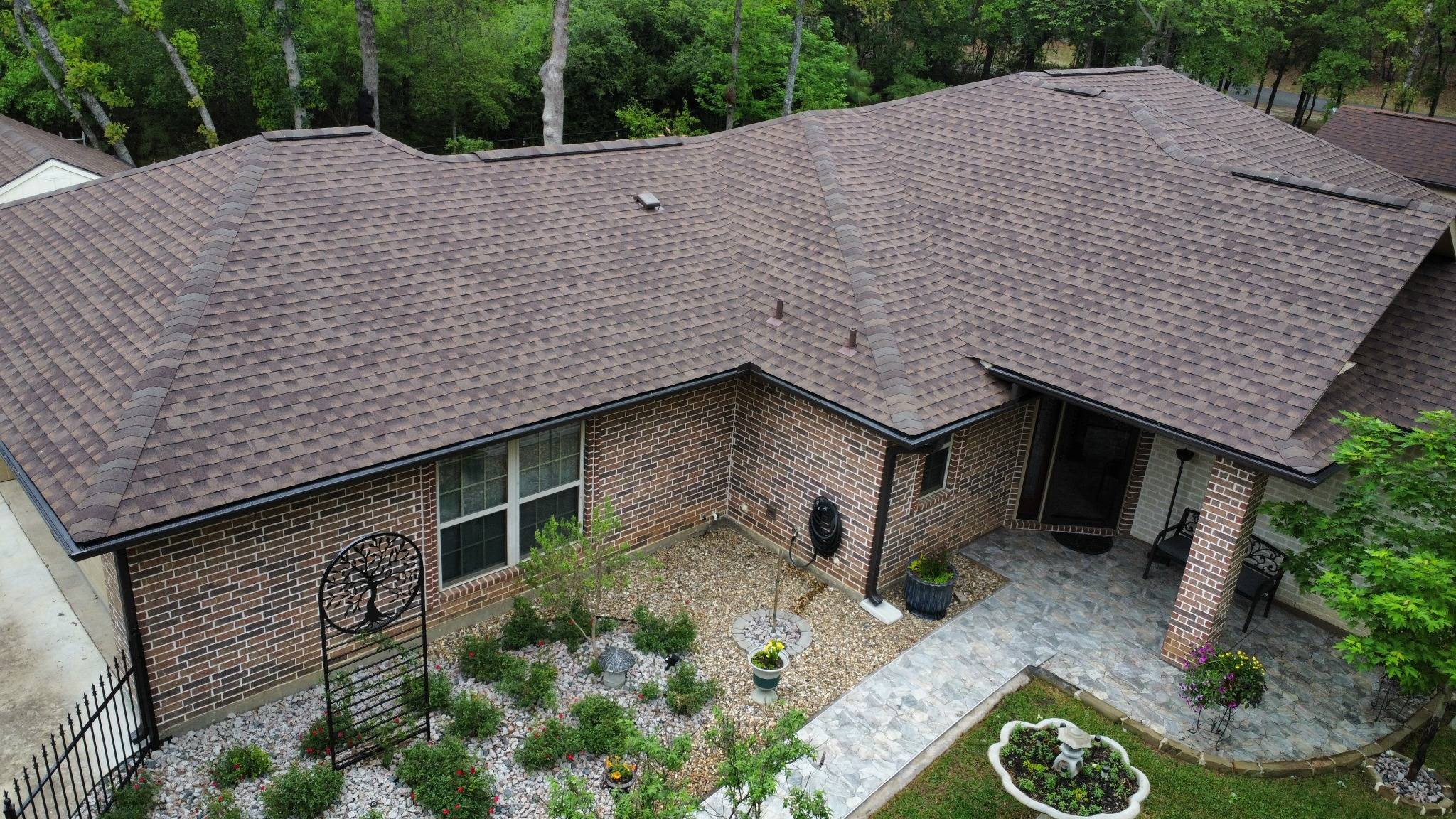A brick home with a brown shingle roof and neatly landscaped front yard surrounded by trees, viewed from above.