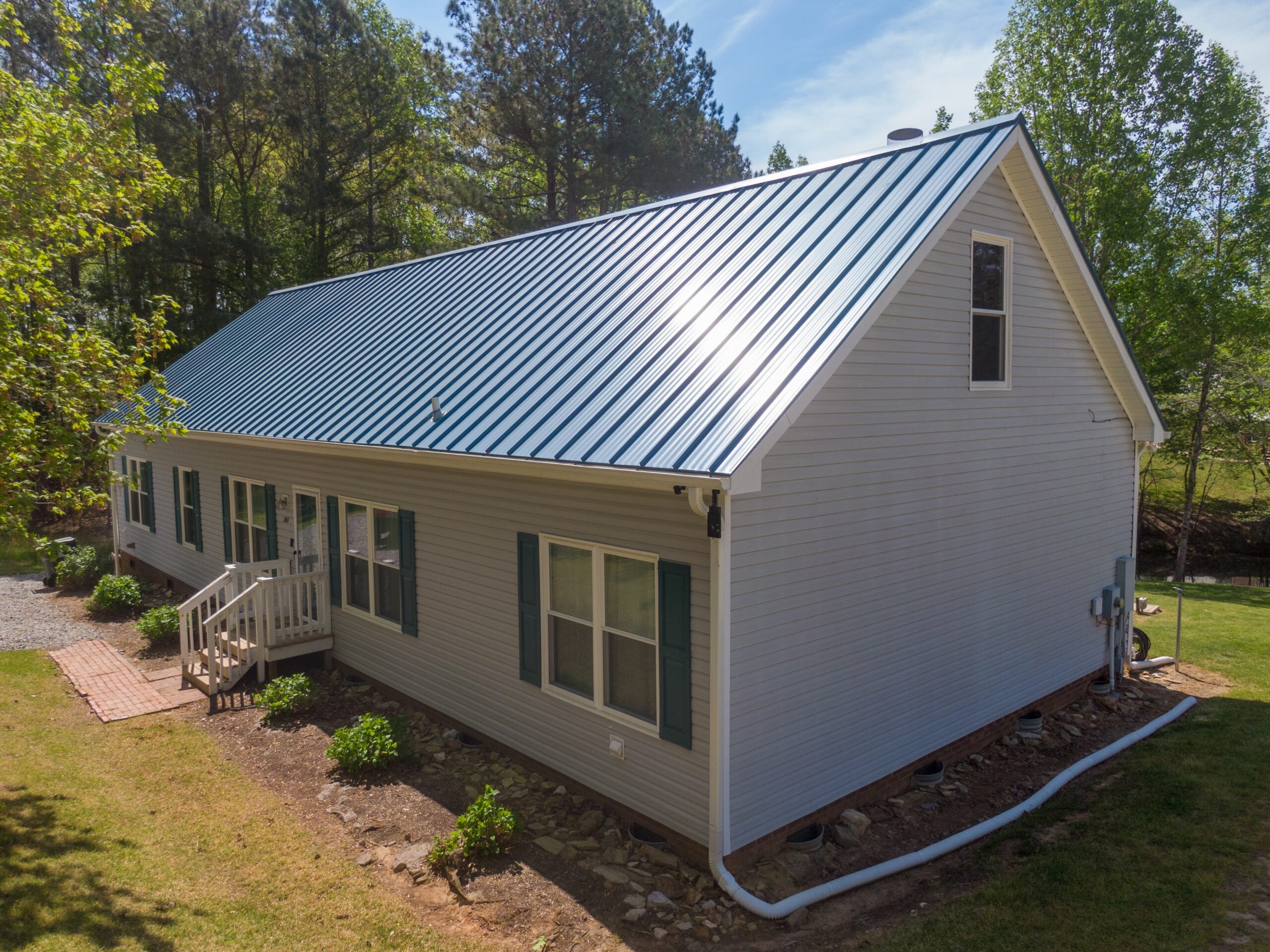 A modern house with light-colored siding features a shiny blue metal roof, surrounded by green trees and a well-kept yard